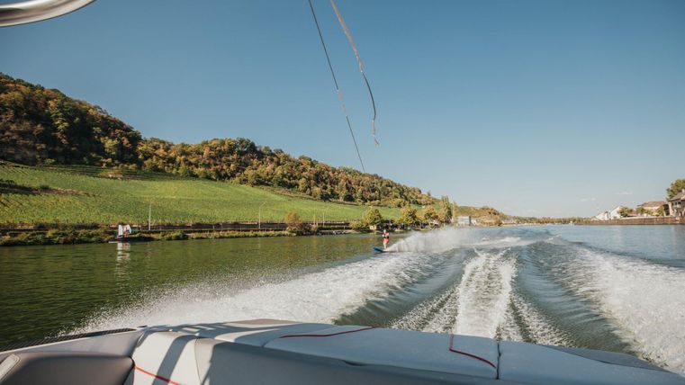 Een boot vaart op een rustig water, omringd door zachte heuvels en groene weiden. De lucht is helder en blauw.