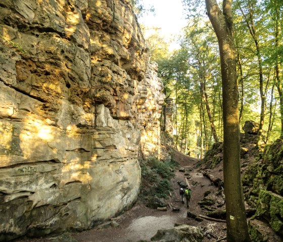 Zwei Wanderer gehen durch die Teufelsschlucht, flankiert von hohen Felsw&auml;nden und umgeben von dichtem Wald im Sonnenlicht., &copy; Eifel Tourismus GmbH, Dominik Ketz