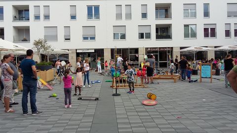 Een drukke speeltuin met kinderen die verschillende activiteiten ondernemen. Omringd door moderne woongebouwen en mensen die toekijken of meedoen.