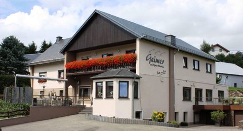 Ein charmantes Gebäude mit blühenden Balkonen und einem modernen Dach. Die Fassade ist hell und einladend, umgeben von grüner Landschaft.