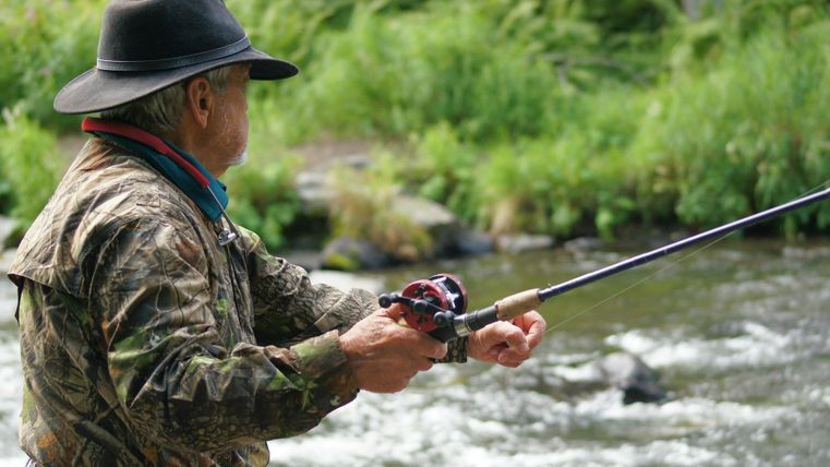 An older man is fishing at the riverbank. He is wearing a camouflage jacket and a hat while he focuses on fishing with the rod.