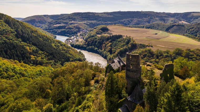 Een schilderachtig landschap met een oude kasteelruïne en een rivier die door de groene valleien stroomt. De omliggende heuvels zijn bedekt met bossen en velden.