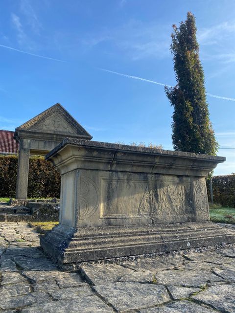 Ein historisches Steingrab mit kunstvollen Verzierungen steht auf einem Pflasterweg. Im Hintergrund ist ein hoher Baum und eine kleine Architektur zu sehen.