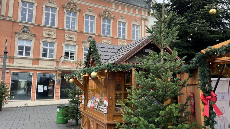 Een feestelijk versierde kerstkraam met kerstbomen in een stad. Op de achtergrond is een historisch gebouw te zien.