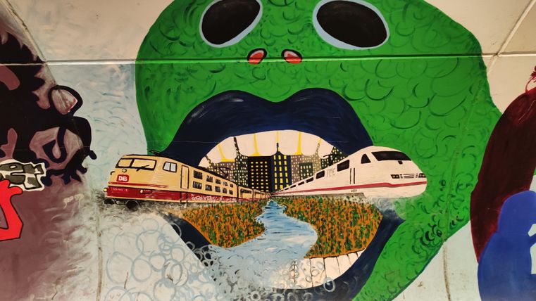 Een kleurrijke muurschildering toont een groot groen hoofd met een open mond. Uit de mond rijden treinen door een stadslandschap, dat een creatieve verbinding tussen natuur en urbaniteit weergeeft.