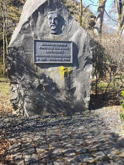 Ein Gedenkstein mit einer Büste und einer Inschrift. Der Stein steht in einem parkähnlichen Umfeld.