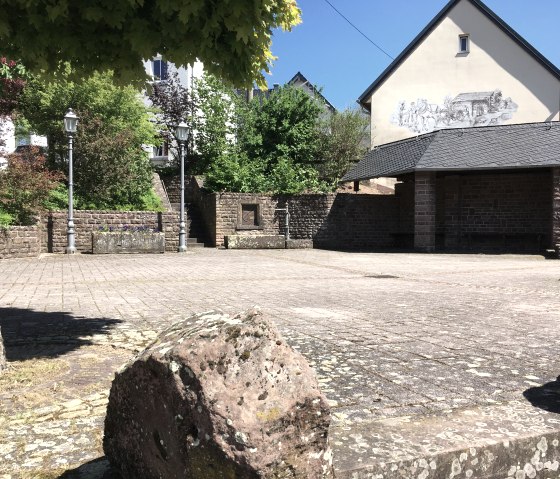 Place de village pav&eacute;e &agrave; Gransdorf avec des murs en pierre, des lanternes et un b&acirc;timent avec des peintures murales. Des arbres et des arbustes entourent la place., &copy; TI Bitburger Land M.Mayer