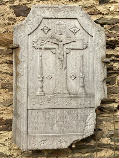 A historical tombstone made of stone featuring a relief cross and a depiction of Jesus Christ. Below, engraved inscriptions can be seen.