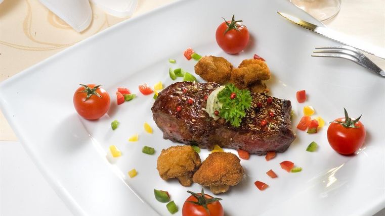 A juicy steak on a white plate, garnished with fresh tomatoes and colorful vegetables. Next to it are crispy sides.