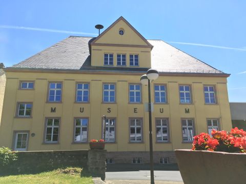 Un bâtiment jaune avec l'inscriptions « MUSÉE » sur le devant. Il a un toit en pente et est entouré de paysages verts et de fleurs.