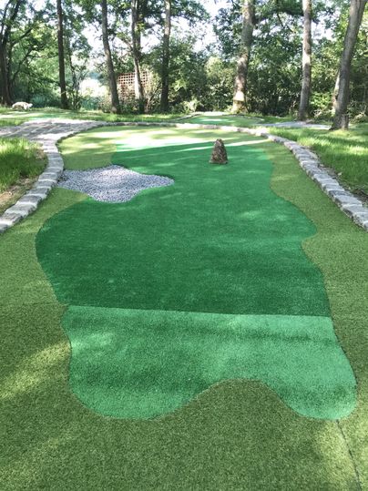 A miniature golf course with artificial grass and a small sand bunker. Surrounded by trees and a paved path.