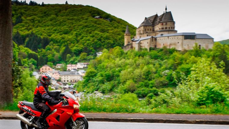 A motorcyclist is riding on a road, while a castle on a hill is visible in the background. The surroundings are green and hilly, with forested areas.