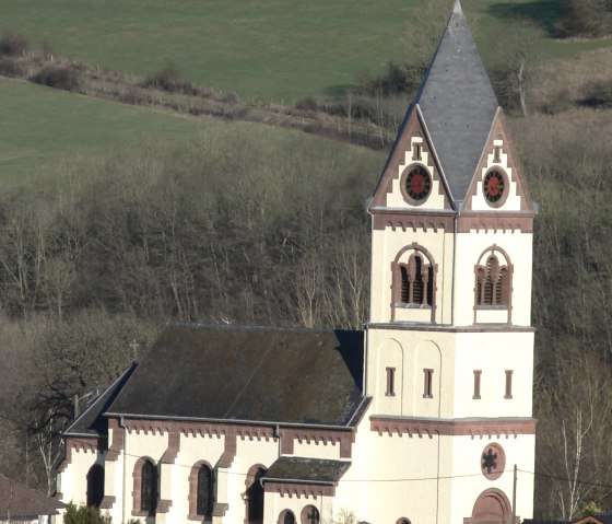 Une &eacute;glise avec une haute tour se dresse dans un paysage rural verdoyant. La tour a un toit pointu et des fen&ecirc;tres d&eacute;coratives., &copy; TI Bitburger Land