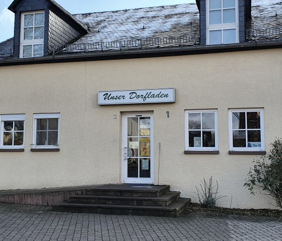 B&acirc;timent jaune avec un panneau 'Notre magasin de village' au-dessus de l'entr&eacute;e. Des pav&eacute;s et un petit buisson devant., &copy; TI Bitburger Land - Steffi Wagner
