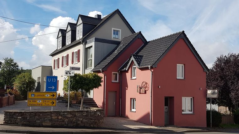 A charming combination of two houses, one pink and one gray, at a street corner. The buildings are surrounded by trees and visible under a clear sky.