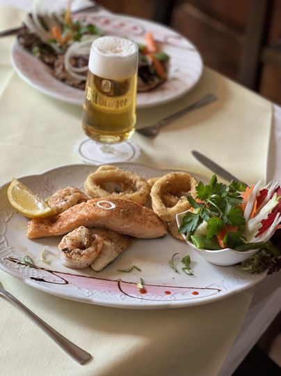 A plate with grilled fish, shrimp, and calamari, garnished with lemon slices and fresh vegetables. In the background, there is a glass of beer.