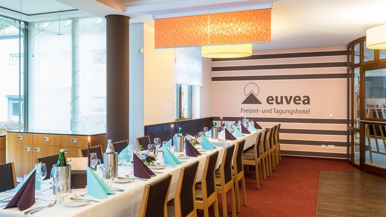 Een elegante, lichte eetzaal met een gedekte tafel en stoelen. De tafels zijn voorbereid met hoogwaardige tafelkleden en decoratie voor een evenement.