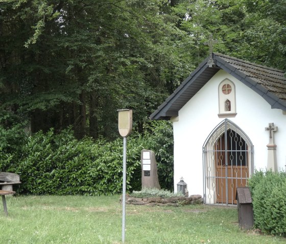 Une petite chapelle blanche avec une croix et une porte grillag&eacute;e se dresse dans la verdure. A c&ocirc;t&eacute;, un banc et un panneau. Entour&eacute; d'arbres et d'arbustes., &copy; TI Bitburger Land