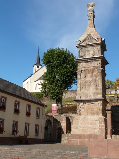 Eine historische Statue steht in der Mitte des Bildes, umgeben von Bäumen und Gebäuden. Im Hintergrund sind sanfte Hügel und ein klarer Himmel zu sehen.