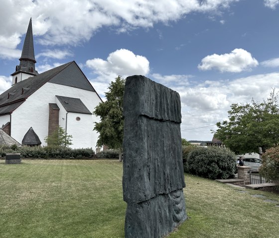 Une église avec une tour pointue à côté d'une sculpture moderne dans le jardin de la bibliothèque Günther Förg à Weidingen., © Felsenland Südeifel, Anna Carina Krebs