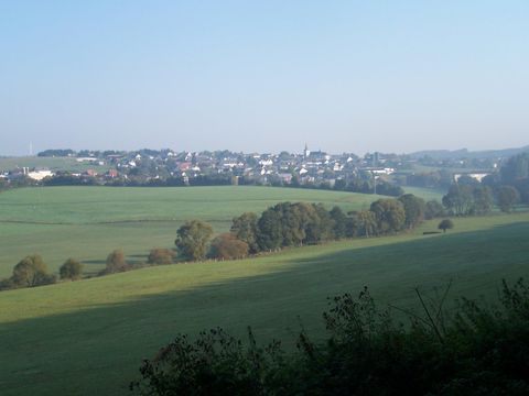 Eine malerische Landschaft mit sanften Hügeln und einer kleinen Stadt im Hintergrund. Der Himmel ist klar und die Wiesen sind grün.