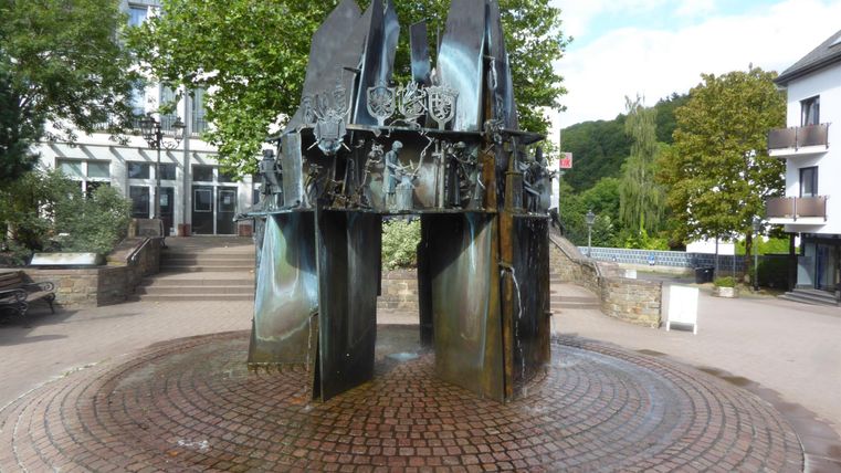 Une installation de fontaine moderne avec des formes métalliques élégantes se trouve au centre d'une place. Entourée d'arbres et de bâtiments, elle dégage un charme particulier.
