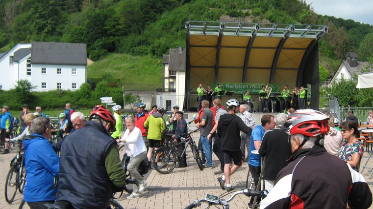 Een groep fietsers verzamelt zich op een plein voor een podium. Op de achtergrond zijn groene heuvels en gebouwen te zien.