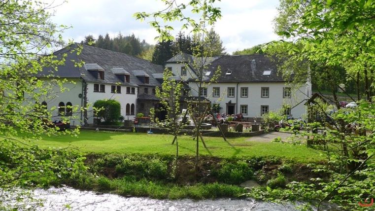 A charming building in a green environment with a river in the foreground. The terrace is furnished with flowers and seating.