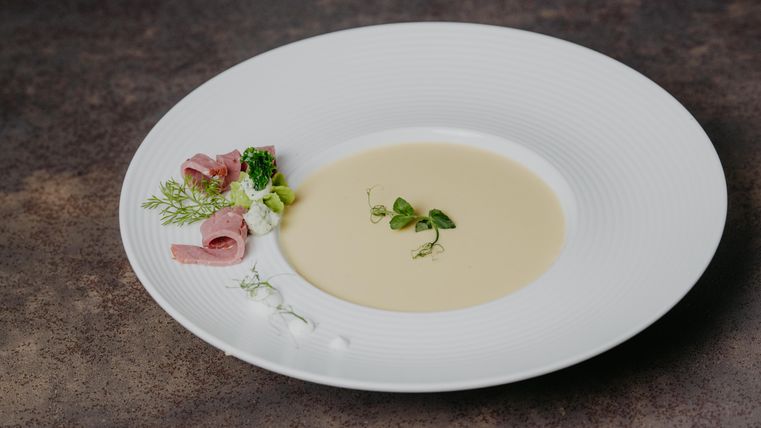 A creamy soup in a white bowl, elegantly arranged. Next to the soup are ham and fresh herbs.