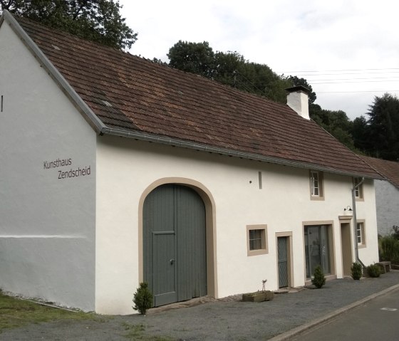 B&acirc;timent blanc avec l'inscription 'Kunsthaus Zendscheid', portail vert, entour&eacute; d'arbres et d'une route. Des personnes sont visibles au premier plan., &copy; R. Schaal
