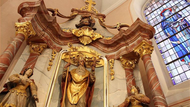 Ein prächtiger Altar mit goldenen Verzierungen und Statuen von Heiligen. Im Hintergrund ist ein bunt verglastes Fenster zu sehen.