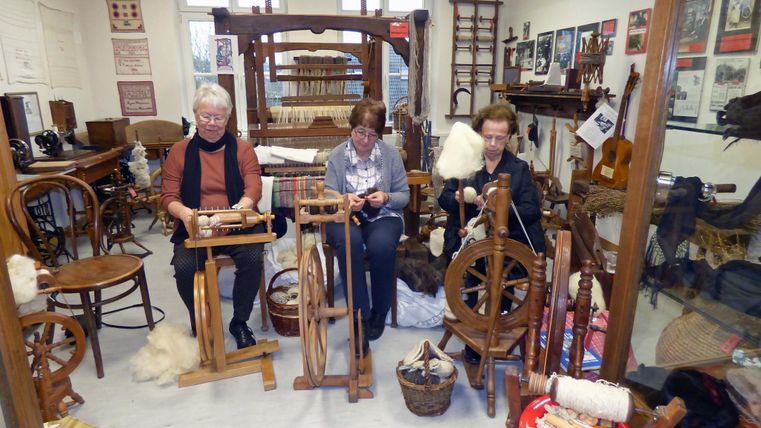 Trois femmes filent de la laine sur des rouets dans un atelier artisanal traditionnel. En arrière-plan, on peut voir divers outils et des images historiques.