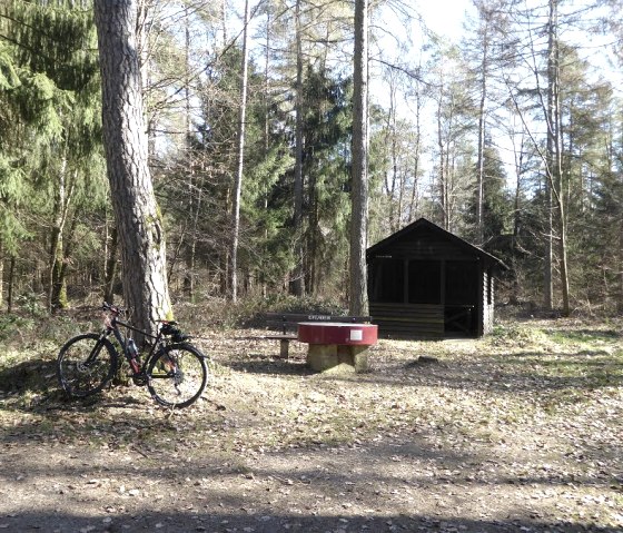 Un v&eacute;lo est adoss&eacute; &agrave; un arbre pr&egrave;s d'un banc dans la for&ecirc;t. En arri&egrave;re-plan se trouve une petite cabane en bois., &copy; Eifelverein Ortsgruppe Speicher