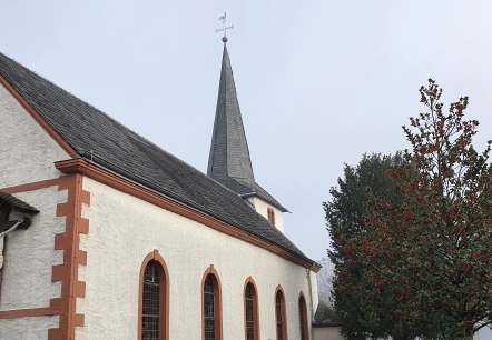 Eine Kirche in Idesheim mit einem Kreuz an der Fassade und einem Baum mit roten Beeren im Vordergrund., &copy; TI Bitburger Land