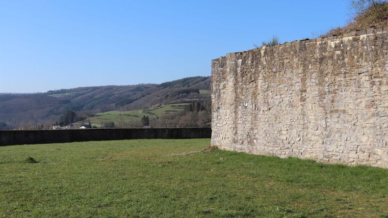 An old stone wall stands in a green field under a clear blue sky. Gentle hills are visible in the background.