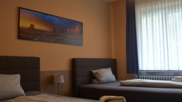 A simple room with two beds and a painting on the wall. There are large windows that let in plenty of daylight.