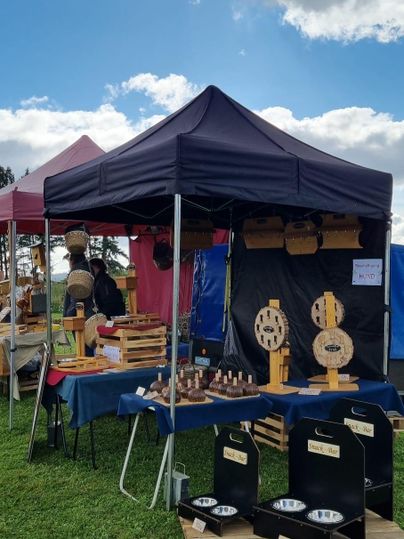 A market stall with a black tent and handcrafted wooden products. In the background, there are more stalls and a blue sky with clouds.