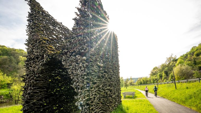 Een kunstzinnige structuur van takken staat aan de rand van de weg. Twee fietsers rijden voorbij terwijl de zon opkomt.