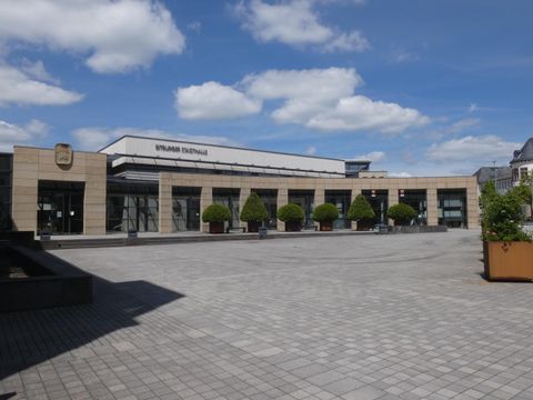 Een modern gebouw met grote ramen en een heldere lucht. Het plein voor het gebouw is bestraat en versierd met groene struiken.