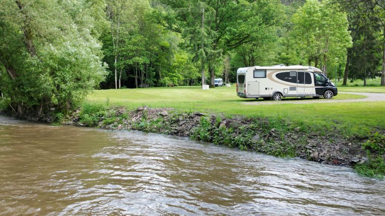 Een camper staat op een weide naast een lichtstromende rivier. Omringd door groene bomen biedt de scène een rustige, natuurlijke sfeer.