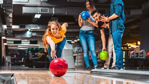 Eine Gruppe von Freunden spielt Bowling in einer modernen Bowlingbahn. Die Frau in der Vordergrund wirft eine rote Bowlingkugel, während ihre Freunde aufmerksam zuschauen.
