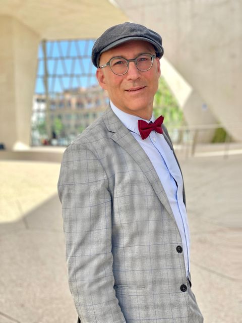 Een stijlvol geklede man in een grijs kostuum met een rode vlindertje en een pet. Hij staat voor een moderne architectuur op de achtergrond.