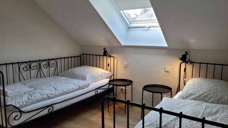 A cozy bedroom with two single beds and window light. The walls are plain and the furniture is made of black metal.