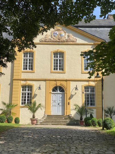 An elegant building with a yellow facade and white windows. The entrance area is surrounded by plants and a paved path.