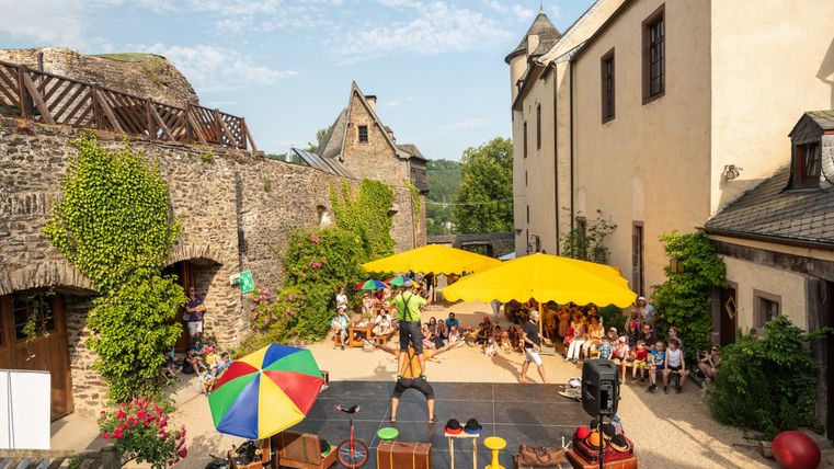 Eine lebhafte Aufführung in einem historischen Innenhof mit bunten Sonnenschirmen. Zuschauer sitzen entspannt auf dem Boden und genießen die Darbietung.