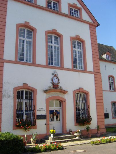A historic building with a white facade and pink details. Large windows and flowers in front of the entrance give it an inviting character.