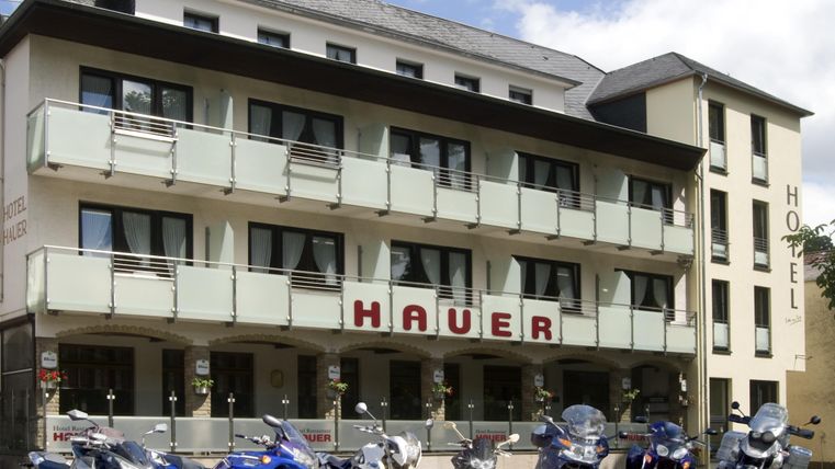 Ein modernes Hotel mit der Aufschrift "HAUER" und mehreren Motorrädern vor dem Eingang. Die Fassade ist hell und einladend, umgeben von einem klaren Himmel.
