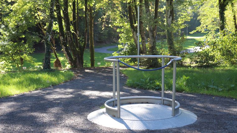 Ein runder Metallring steht auf einem gepflasterten Weg in einem grünen Park. Umgeben von Bäumen und Wiese bietet die Szene eine ruhige, natürliche Atmosphäre.