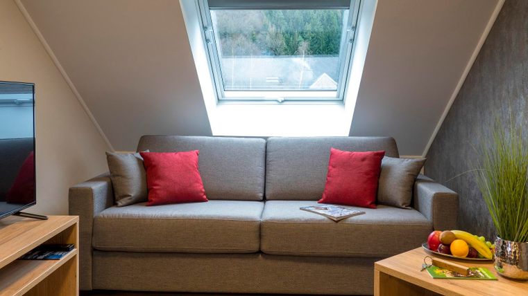 Ein modernes Wohnzimmer unter dem Dach mit einem bequemen grauen Sofa und roten Kissen. Der Raum ist hell erleuchtet und verfügt über ein großes Fenster sowie eine gemütliche Atmosphäre.