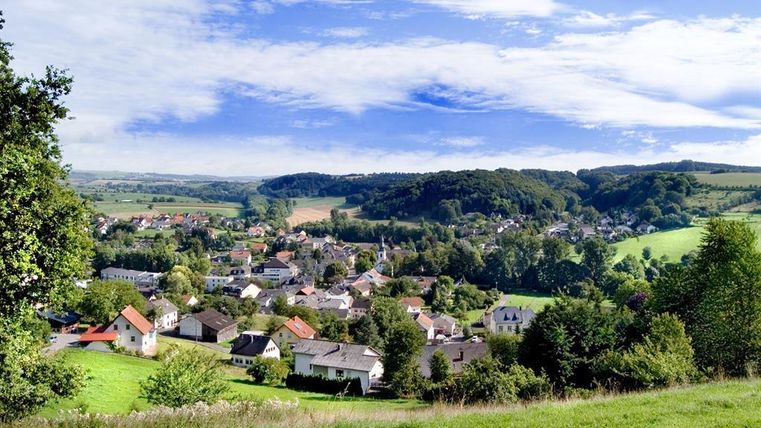 A picturesque landscape with a small village nestled between gentle hills. The sky is clear and nature is green and inviting.